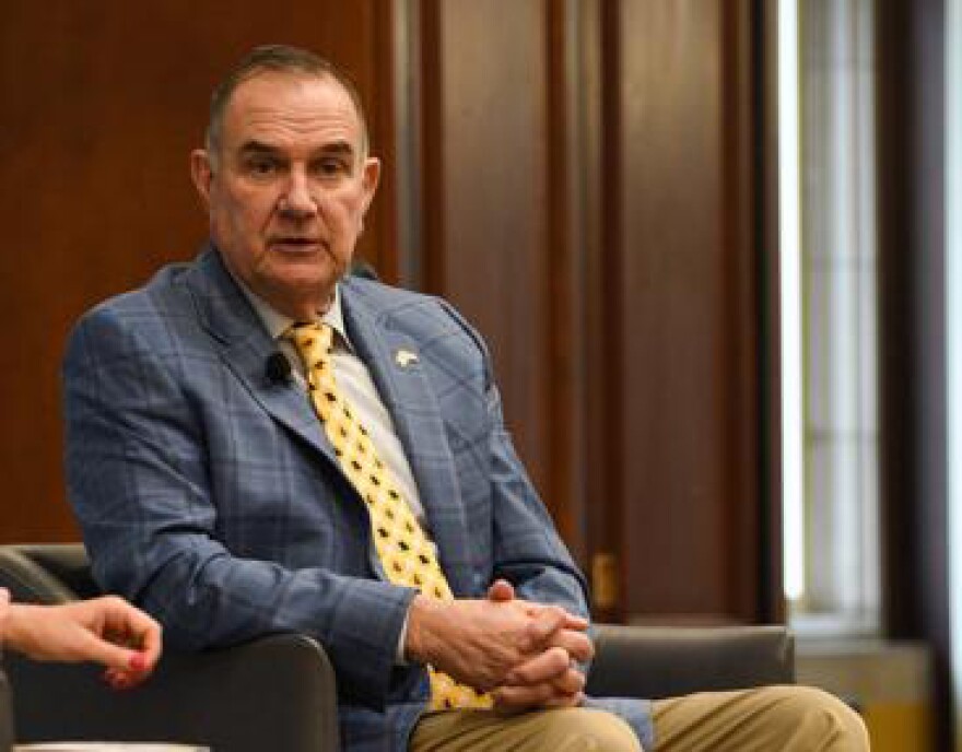 Governor Mike Kehoe sits in a chair located on the University of Missouri campus. He is wearing a pale blue jacket and yellow tie and is speaking with his hands folded on his knee.