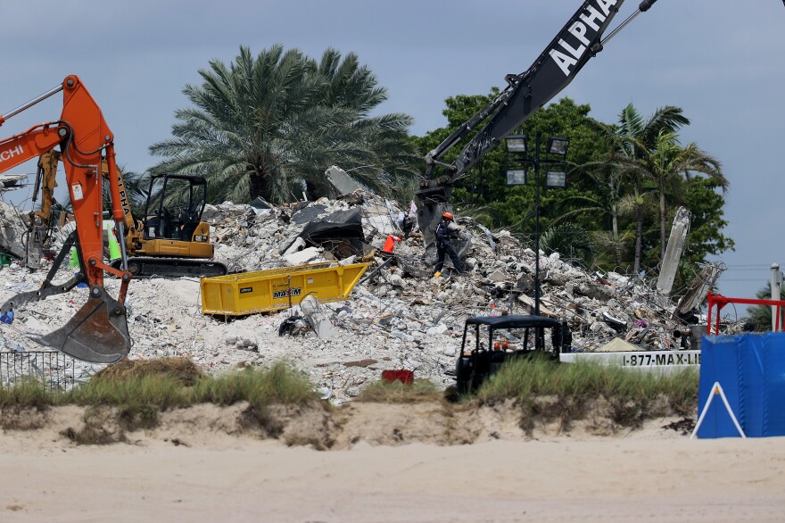 Crews and a bulldozer sift through rubble, with a sandy beach in the foreground and green palm trees behind it.