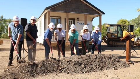 Members or Watered Gardens Ministries break ground at Freedom Village on September 4. They're wearing hard hats and are holding shovels in front of a pile of dirt/ They're standing in front of a model tiny home.