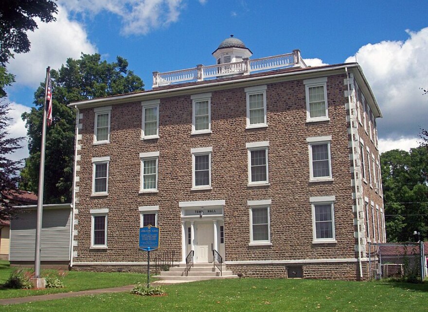 Alexander Town Hall is a three-story cobblestone building with white doors and trim. There's an American flag on the pole on the left side of the image. 