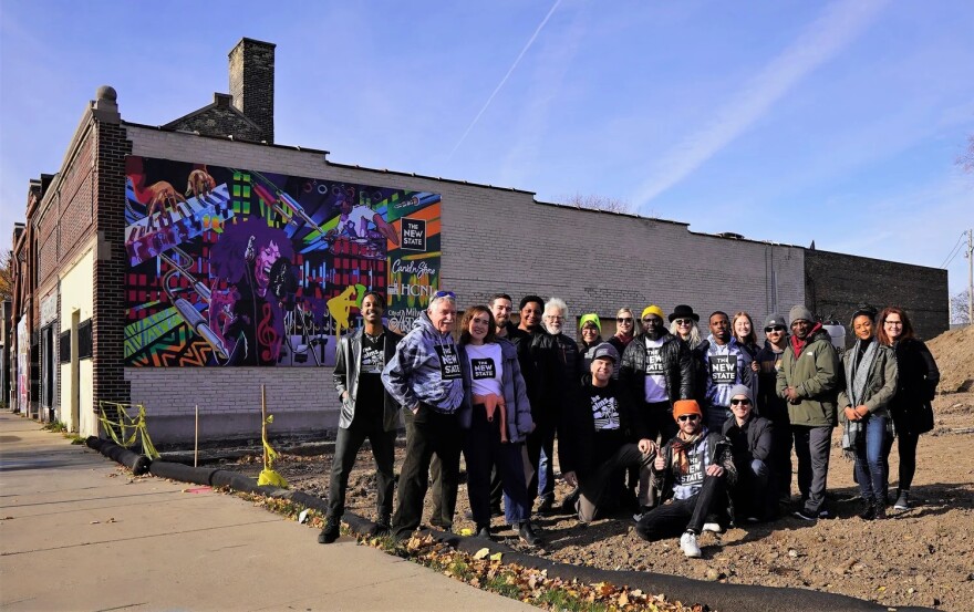 The New State team poses as a group in front of a mural on its building by artist Brad Bernard.