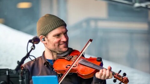 A man wearing a knit hat and button up shirt plays a fiddle in front of a music stand and microphone.