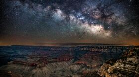 Photo over the Grand Canyon at night. The sky is bright with stars and the Milky Way is visible.