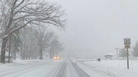 Headlights of oncoming traffic cut through the snow on a road where the pavement is only showing through tire tracks in the snow. 