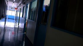 A hallway is empty with most of the lights off and one classroom light still on on what would otherwise be a blended learning school day at Yung Wing School P.S. 124 in New York City.