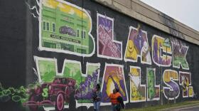 A large, colorful mural reads "Black Wall St." with images of storefronts, buildings, old-time cars, jazz musicians in the letters. Two women are in front of the mural and dwarfed by the size of it.