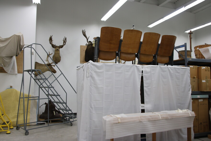 Four wooden movie theater chairs sit on top of shelves covered with white sheets in a basement. In front of the shelves is a metal moving staircase and a taxidermy deer on the wall.