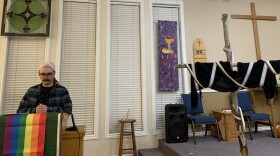 A person in a flannel and trans-themed head wrap stands at a church pulpit decorated with the gay flag. A nearby wooden cross is draped with a black scarf and the trans and nonbinary flags.