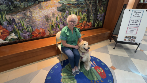 A woman wearing a green top and jeans holds her golden retriever while sitting on a chair in a library lobby.
