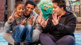 A father and mother are counting on their fingers with their daughter. A green puppet is with them.  