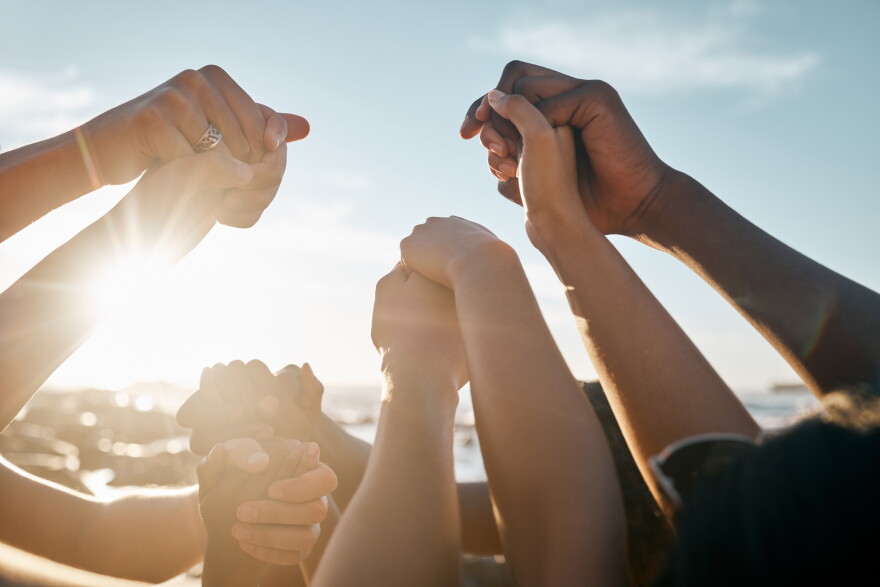A diverse group of people holding hands in the sunshine. Only their forearms and hands are visible.