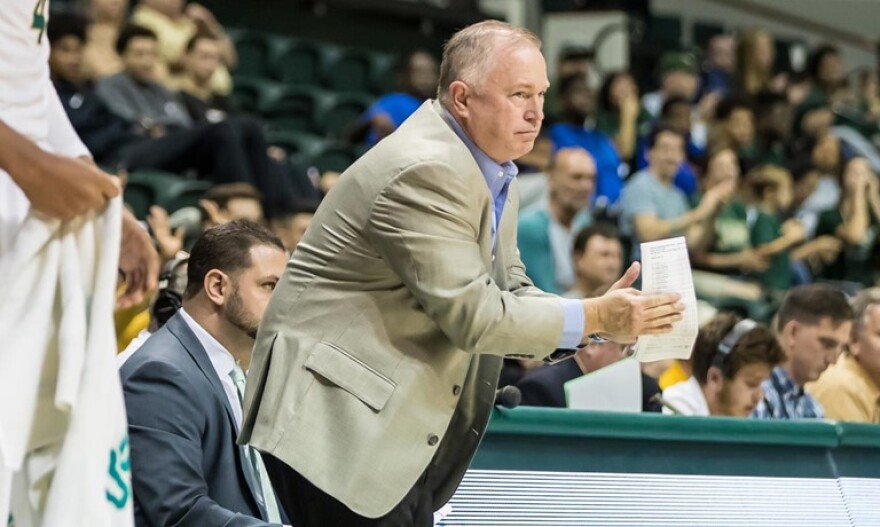 Man in jacket claps while leaning over on the sideline of a basketball game