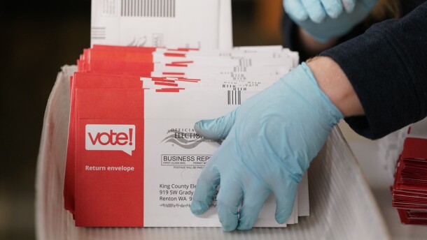 A person holds an orange and white ballot envelope while wearing blue latex gloves. 