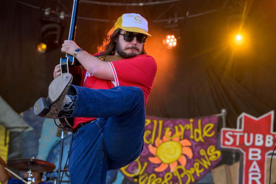 A long-haired bearded man in a trucker hat, cowboy boots, jeans and sunglasses on stage, kicking his leg up and strumming his electric guitar off to his side