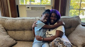 A woman in leopard print pants sits on a tan couch and hugs her daughter, who wears a blue shirt and jeans. 