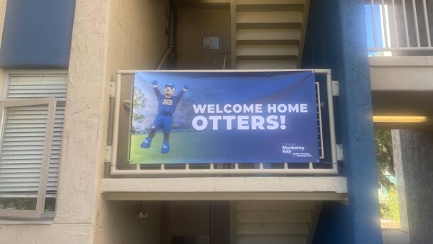 A blue banner with white letters that read "WELCOME HOME OTTERS" on the right of the banner is paired with an image of a college otter mascot standing up with hands raised on the left of the banner. The banner hangs on rails outside a staircase in a college dormitory setting. 