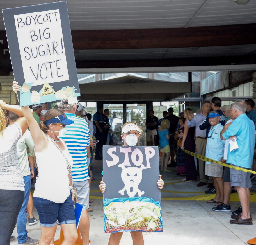 Cape Coral resident S'iva Goodman hoists a sign outside of the Cape Coral Yacht Club beside her 11-year-old daughter, Shelby, who is also carrying a sign protesting the U.S. Army Corps of Engineers and symbolically wearing a surgical mask.