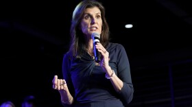 A woman holds a microphone and speaks in front of a darkened background