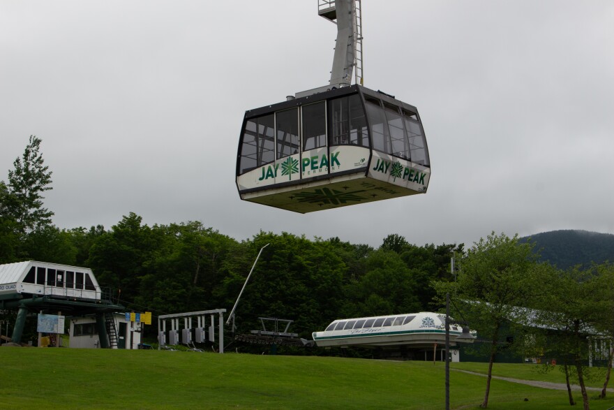 A tram with windows on all sides and text that says "Jay Peak" on the bottom half is suspended in the air, with grass and ski lift equipment in the background.