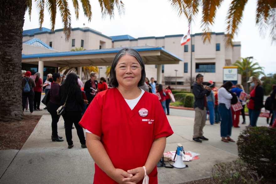 A nurse wearing red scrubs stands in front of Watsonville Community Hospital. Nurses behind her talk to one another while holding protest signs.