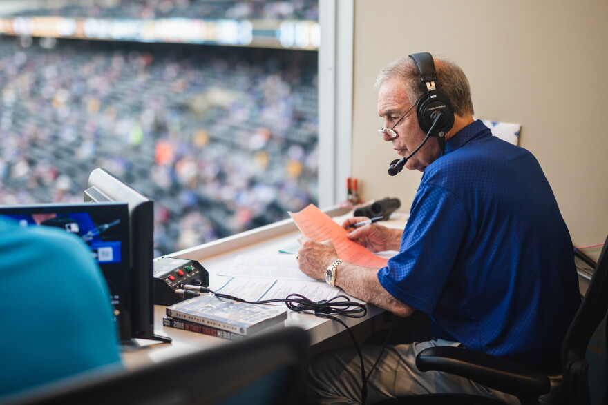 Denny in the booth announcing a game from a press box. He's looking left over the field while wearing a blue Royals polo.
