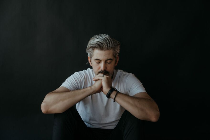 A man with gray hair and a beard sits with his elbows on his knees, hands clasped in front of his face, eyes closed, appearing thoughtful or contemplative, against a dark background.
