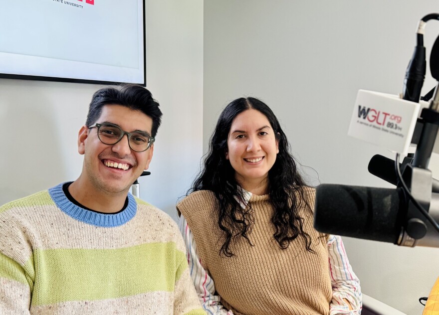 Two people smile while sitting in front of microphones in a radio studio. One person wears glasses and a light-colored sweater with green stripes, and the other has long dark hair and a tan vest over a striped shirt.