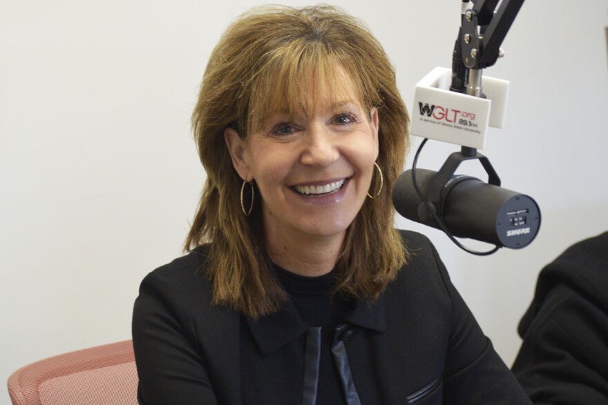 Colleen Kannaday smiling in a radio studio wearing a dark suit and seated next to a microphone with a flag that reads 'WGLT.org 89.1 FM'