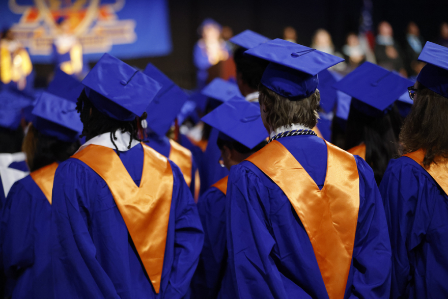 The backs of a crowd of graduates in blue graduation gowns wearing gold sashes