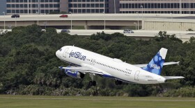 A large, blue and white JetBlue Airways Airbustakes off from the Tampa International Airport. It has just left the ground at a tilted angle and the airport terminals can be seen in the background.