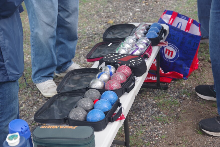 Open bags of boules lie on a table. The boules are different colors - blue, silver with blue stripe, faded red, charcoal. People stand around them