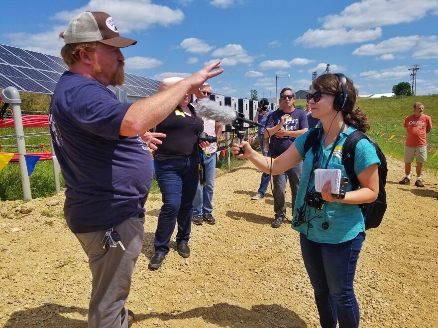 Hope Kirwan stands outdoors in front of a field of solar panels. She wears headphones and a backpack and she's holding a small notebook. She is pointing a mic at a man with a beard and a cap.