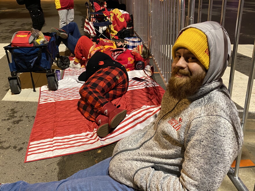 A man in a grey jacket and blue jeans sits on the ground with his back to a temporary metal fence. In the background, people decked out in red and yellow clothing sleep on blankets. 