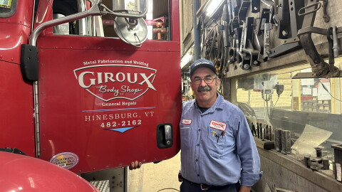 Smiling man in blue workshirt standing by big red red truck which on the door reads 'Giroux Body Shop, Hinesburg, Vermont' 