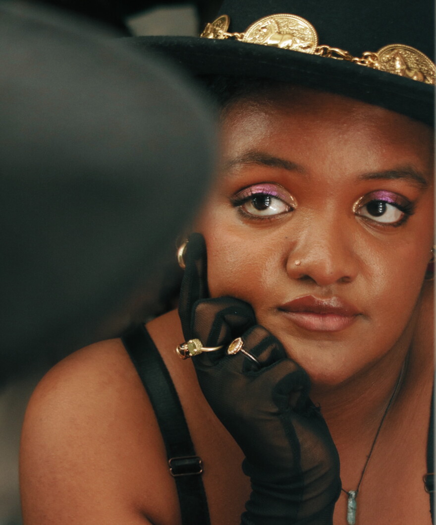 Woman with a black gloved hand on her face and purple eyeshadow looking off camera.
