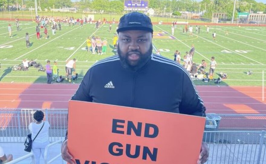 A man wearing a black baseball hat and black Adidas jacket holding an orange sign that says, "END GUN VIOLENCE." He is standing in bleachers in front of a football field. 