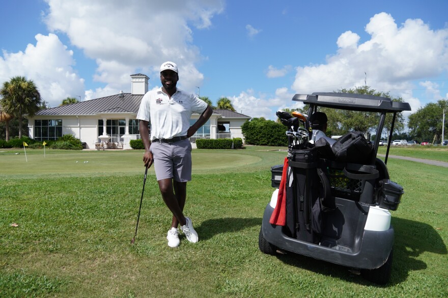 African American man stands with his club on the ground next to a golf cart in front of a clubhouse. 