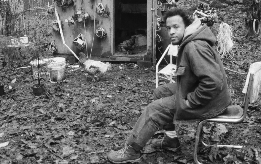 A man sits in a beat-up metal chair in a forest clearing and looks at the camera with a damaged shed in the background. 