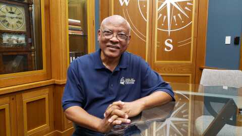 A smiling Black man is sitting at a glass table in a map library with his hands folded  