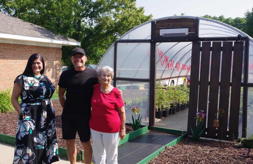 Two women stand with a man in front of a greenhouse filled with potted plants. From left to right, one woman wears a black dress with white and blue flowers decorating it, the man wears all black from his hat to his shorts, the last woman wears a bright red blouse and white pants. 