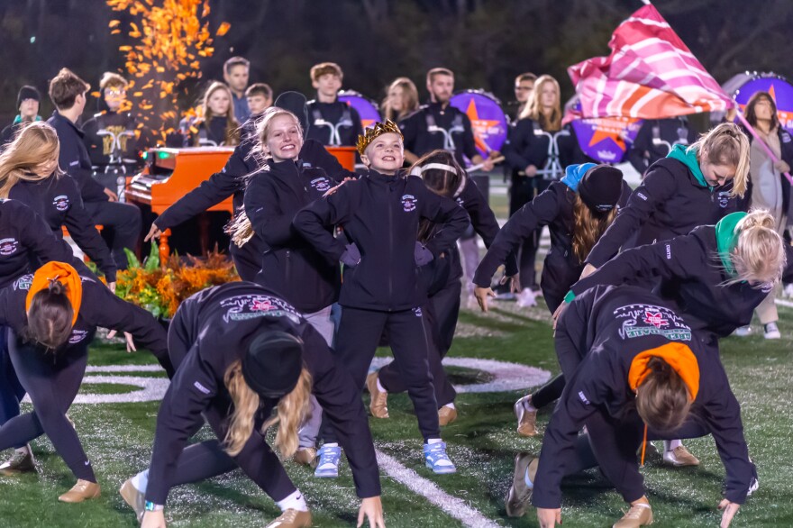 A group of high school students dance on a marching band field. One of them stands in the center of a block formation, and they are wearing a crown.