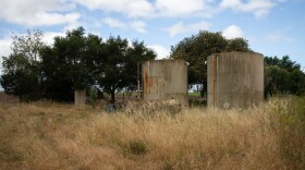 photo shows two abandoned wells in a small grassy field with trees in the background. 