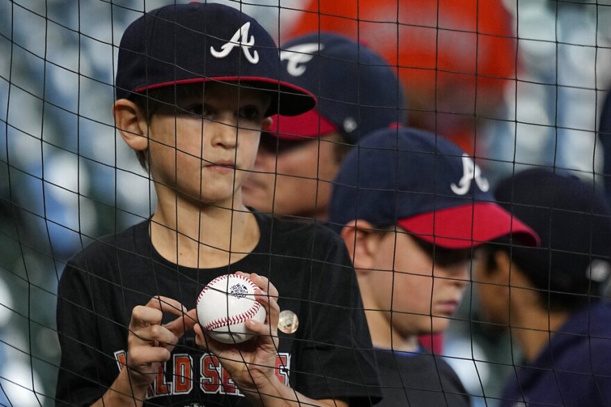 A young boy wearing an Atlanta Braves hat (dark blue with a white script A) holds a baseball behind the netting in front of the stands at a baseball stadium.