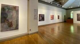 Five rusty colored paintings are hung on white gallery walls with a gleaming maple floor and the word "home" painted vertically on a pillar.