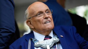 An older, balding man in a suit and glasses with a device around his neck.