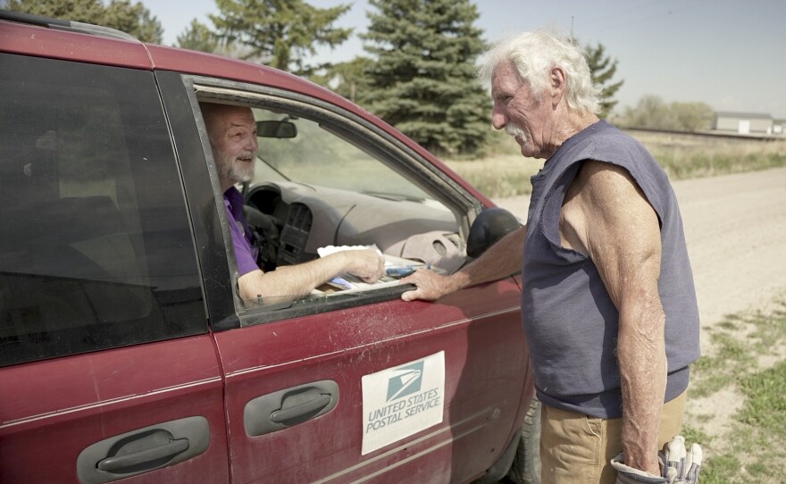 A man with a salt-and-pepper beard smiles from a dusty, red van in rural Gering, Nebraska. On the van's side is a white rectangle that says, "United States Postal Service." The man, Roger McDonald, is talking to another man, Edgar Clemens, who has white hair and a white mustache. A gravel road stretches in front of them. The two men are chatting on May 9, 2025 — McDonald’s first day back on the route after three months of medical leave. McDonald said he loves the sense of community fostered by the Postal Service, which he fears would disappear if his route was curtailed by cutbacks or eliminated by privatization.