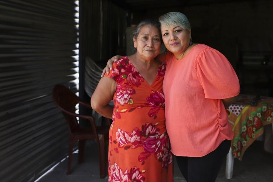 Two women pose for a portrait. They are hugging at the shoulders inside a darkened house.