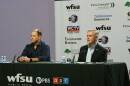Two men sit at a table in front of microphones staring forward.
