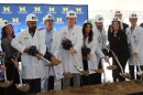 A group of people are lined up wearing white lab coats and hard hats with University of Michigan branding. They are all smiling while holding shovels, digging up a mound of dirt to symbolically break ground for the construction project.