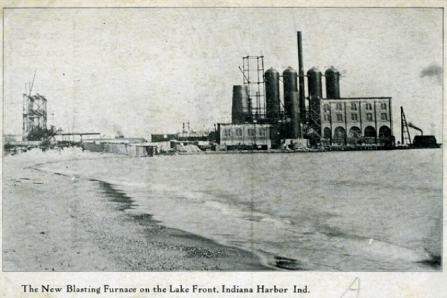 A vintage black and white postcard shows a large industrial complex with several tall smokestacks and cylindrical structures on the waterfront. The foreground is a body of water with a shoreline. The text at the bottom of the image reads, "The New Blasting Furnace on the Lake Front, Indiana Harbor Ind."